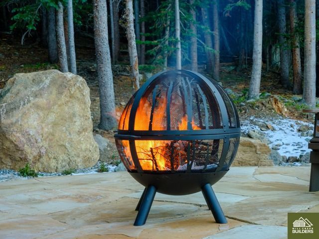 Black spherical fire pit with visible flames burning outdoors on a patio near rocks and trees