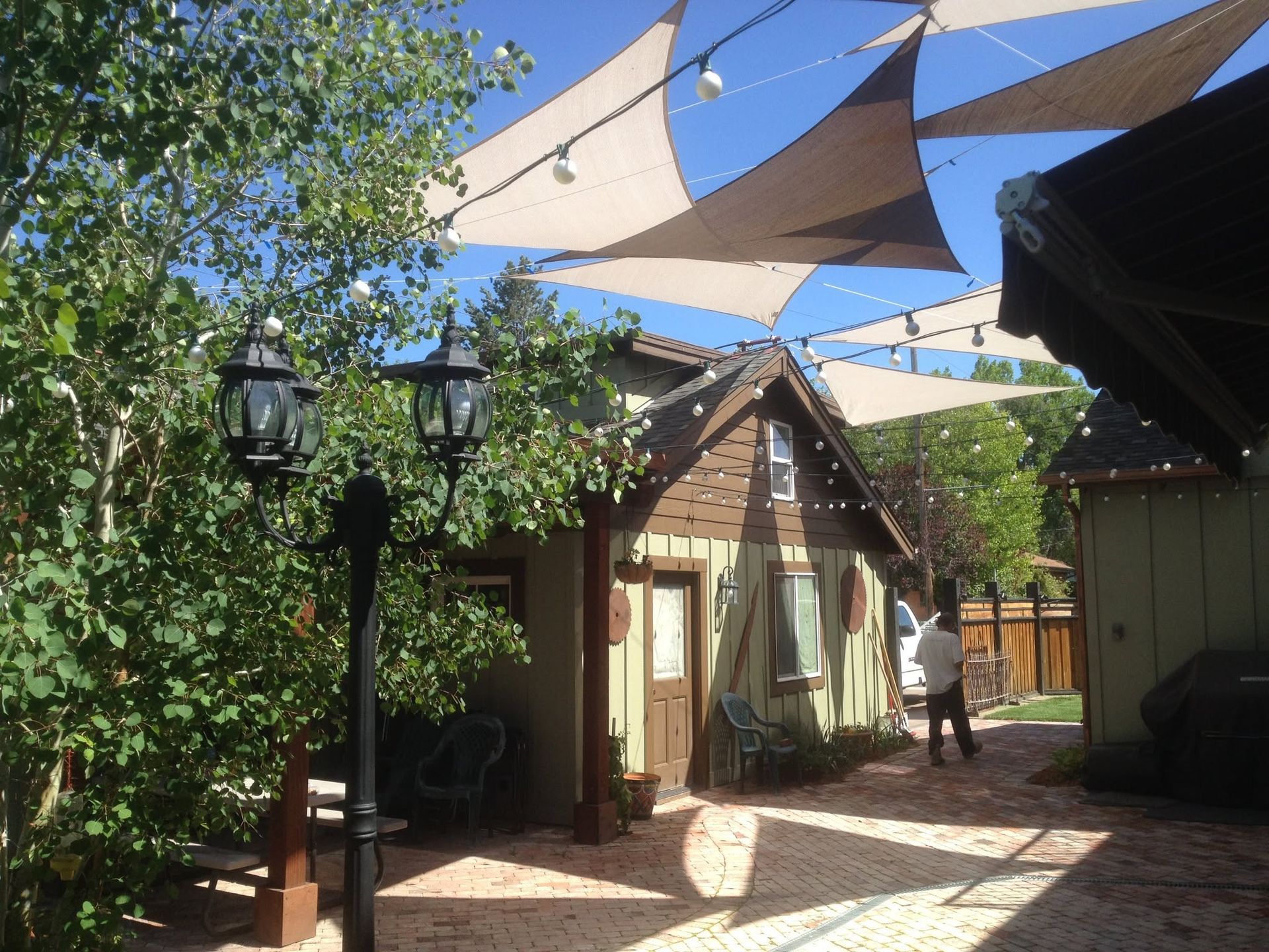Shaded courtyard with a small wooden cottage, leafy trees, and hanging sail canopies under a blue sky