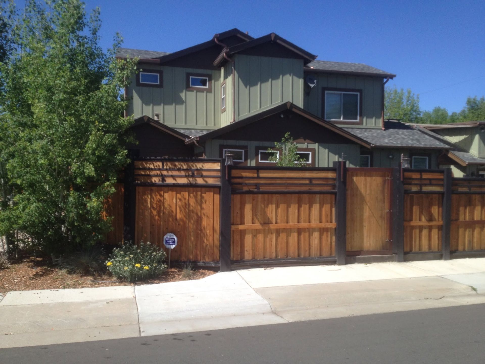 Modern two-story house with green siding and a wooden gate, viewed from the street under a clear blue sky