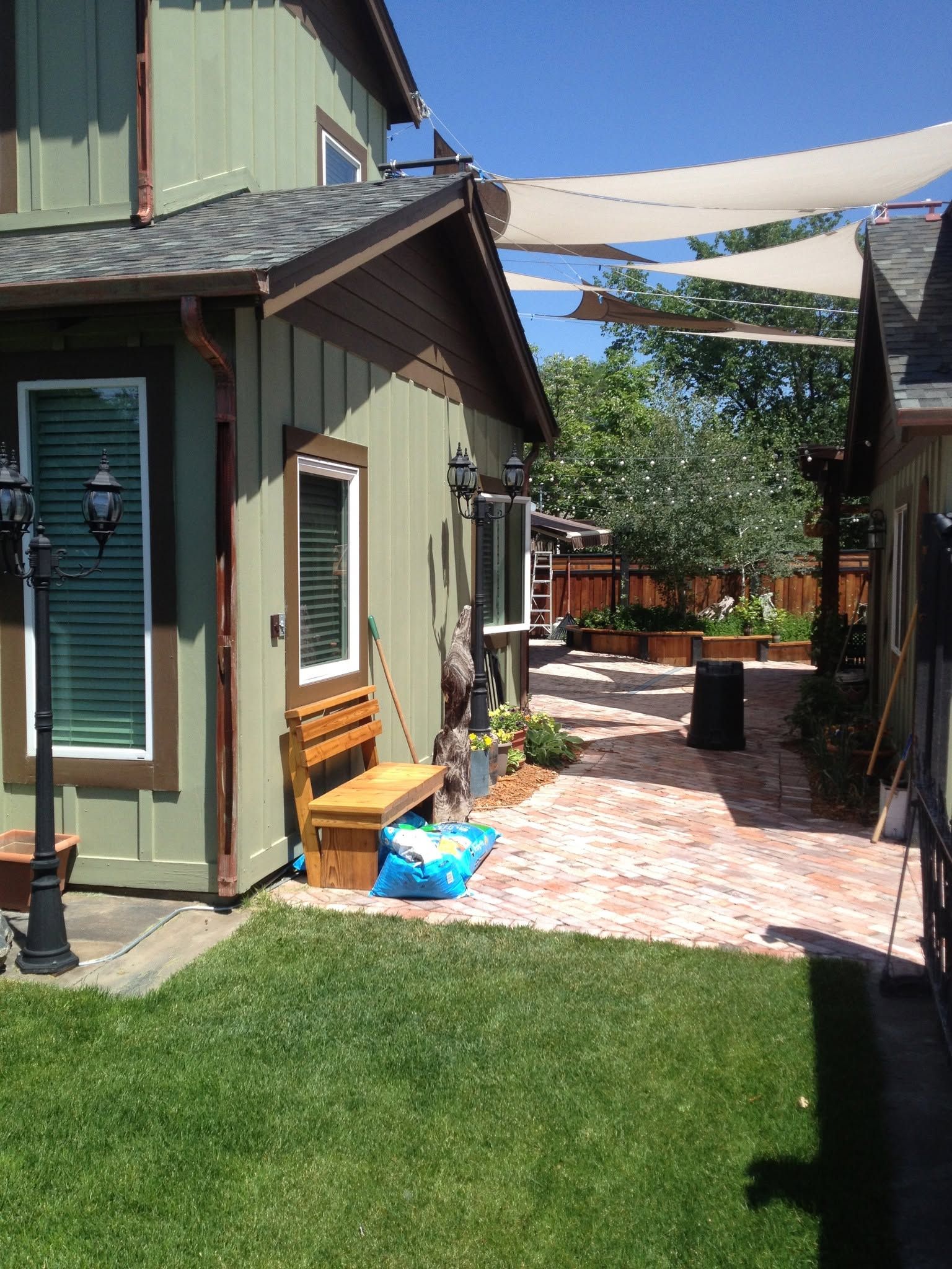 Backyard patio beside a green house with brick walkway, shade sails, and grass lawn