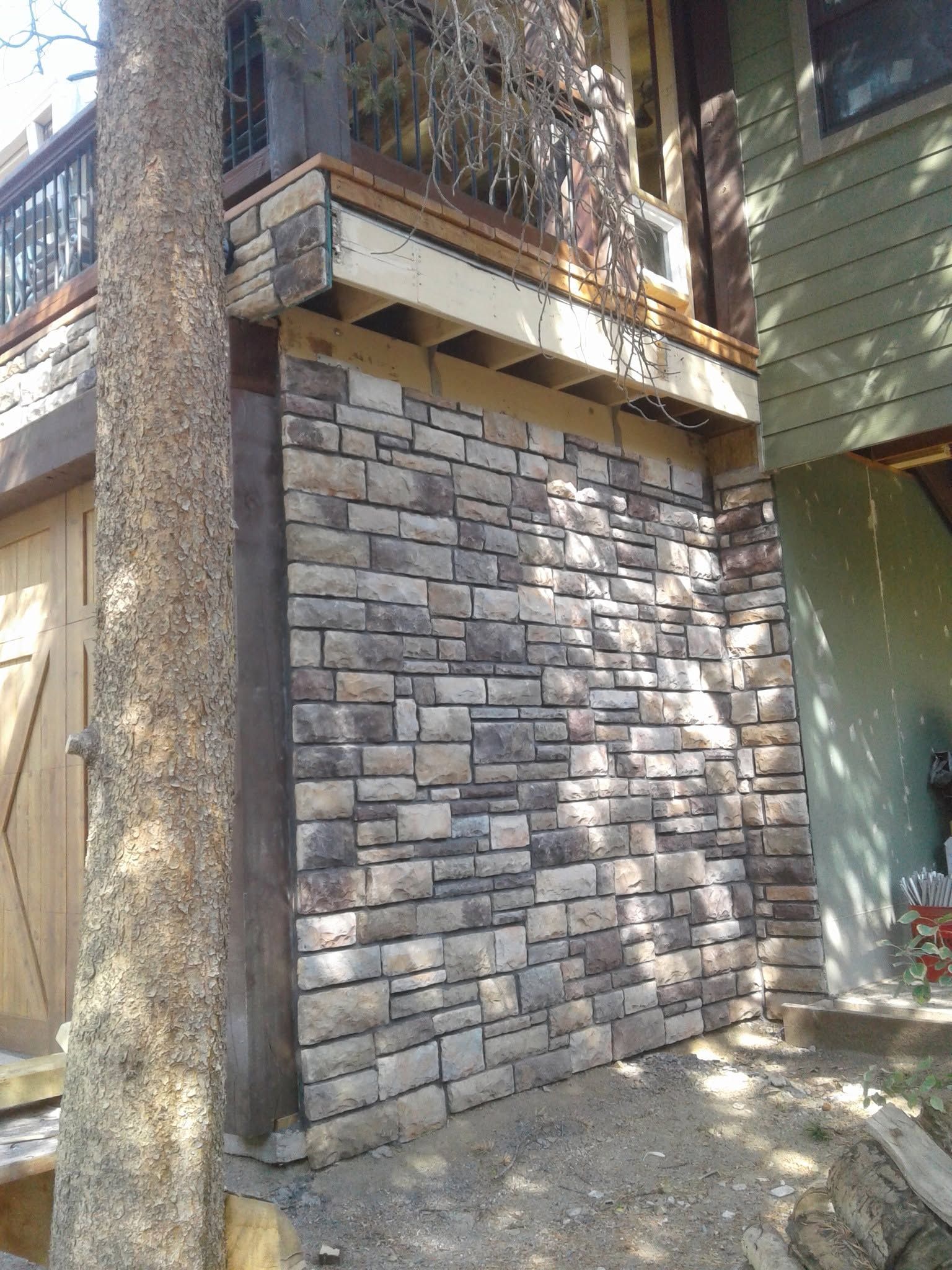 Stone-clad exterior wall beside a house, with a tree trunk in front and a wooden balcony above