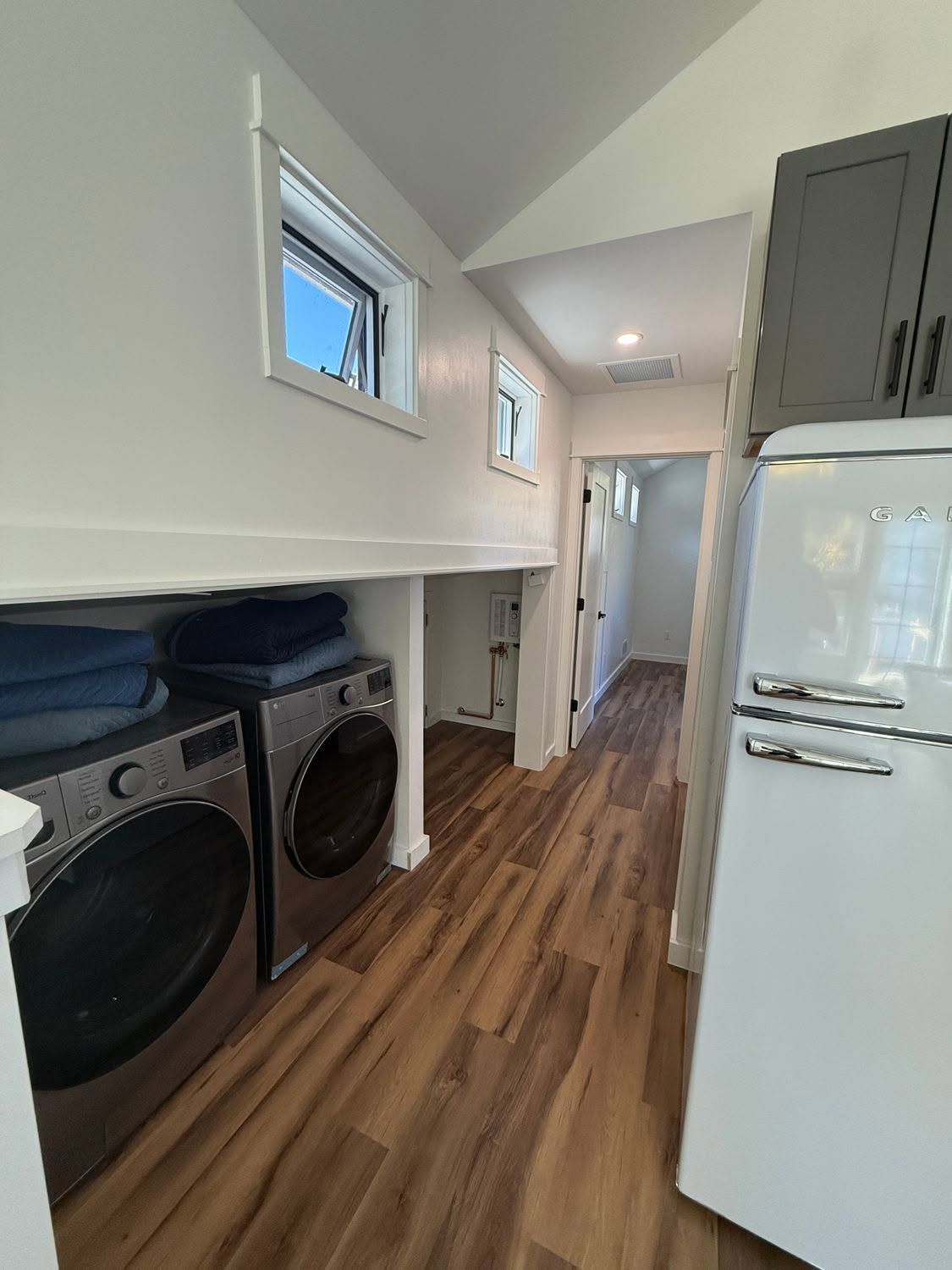 Narrow laundry hallway with washer, dryer, white cabinets, and a refrigerator along a wood floor corridor.