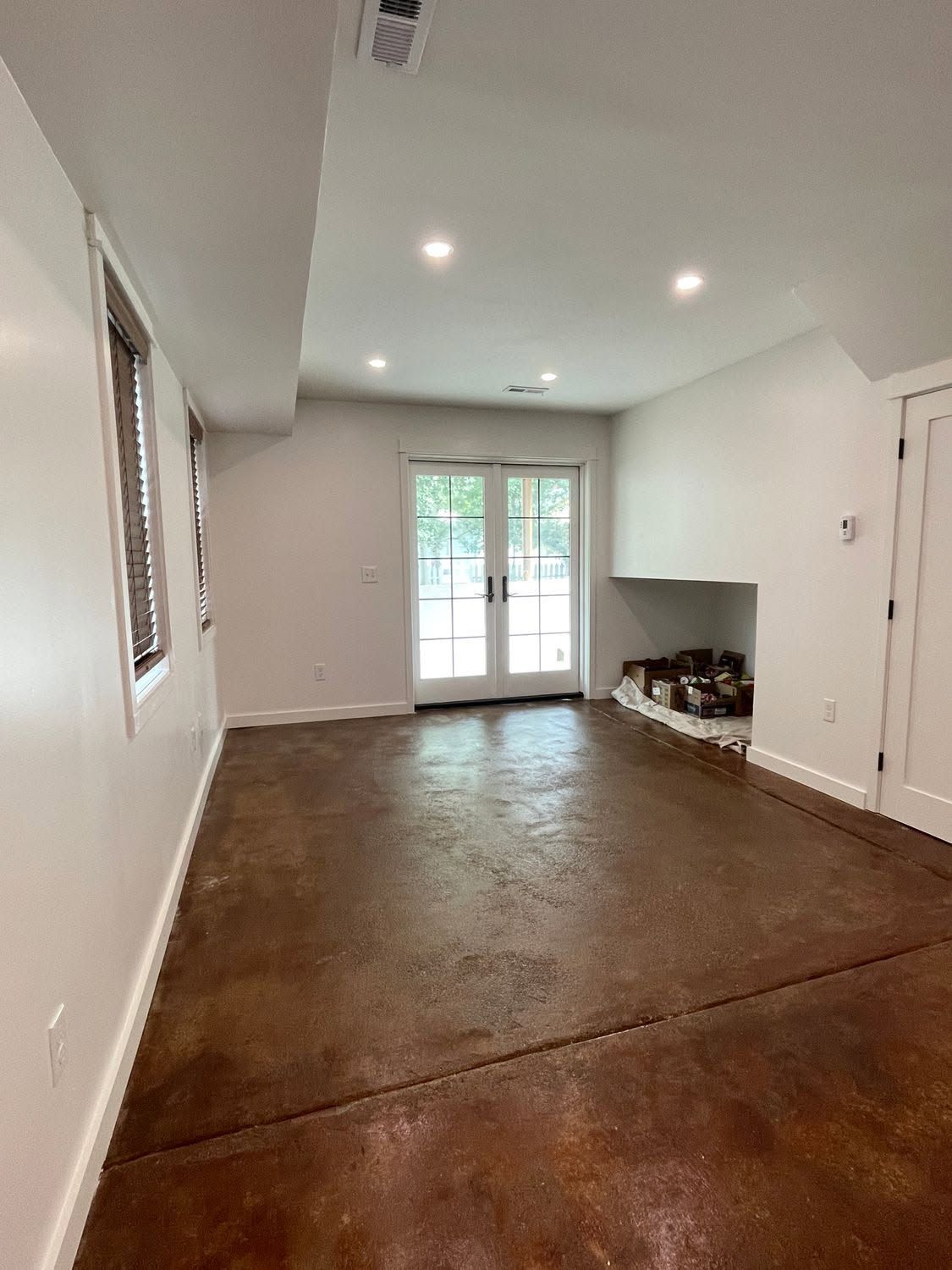 Empty living room with brown floor, white walls, recessed lights, and double doors to a patio.