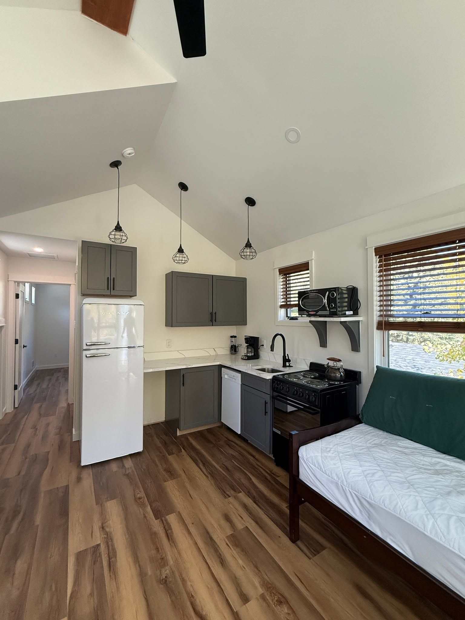 Small studio apartment with kitchenette, bed, white fridge, wood floors, and windows with blinds.