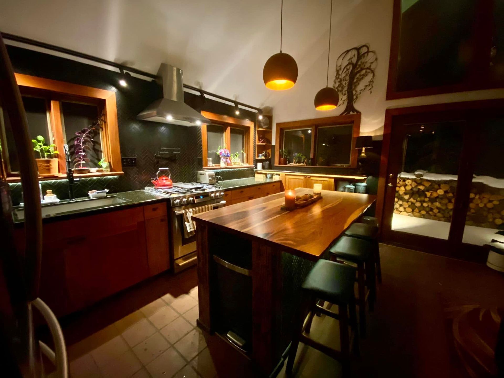 Warm modern kitchen with wooden island, pendant lights, dark cabinets, and tiled floor at night
