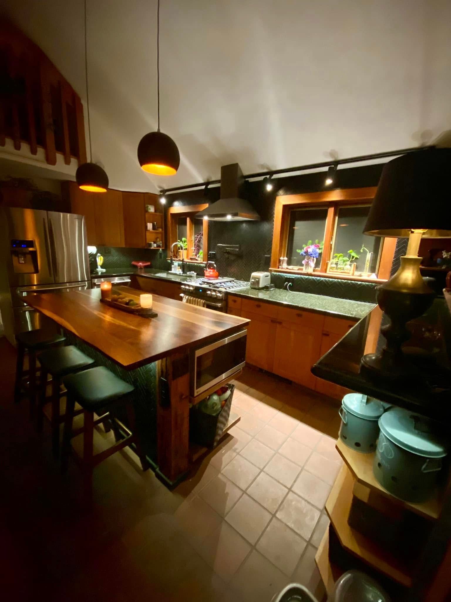 Warm, dimly lit kitchen with wooden cabinets, a central island, bar stools, and copper pendant lights.