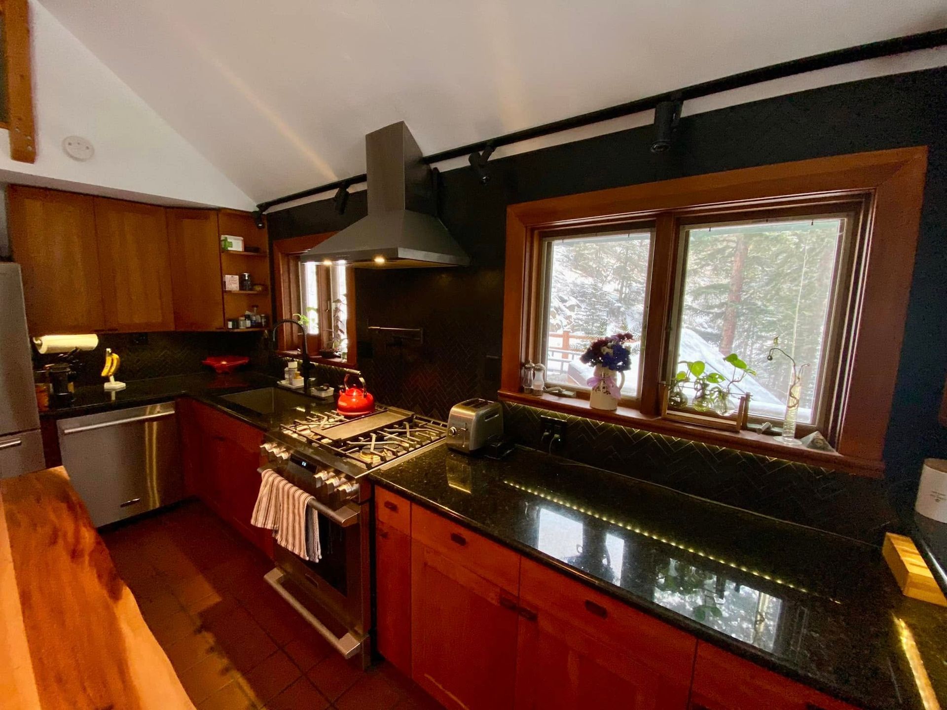 Modern kitchen with wood cabinets, black countertops, stainless stove, and large window over the sink.