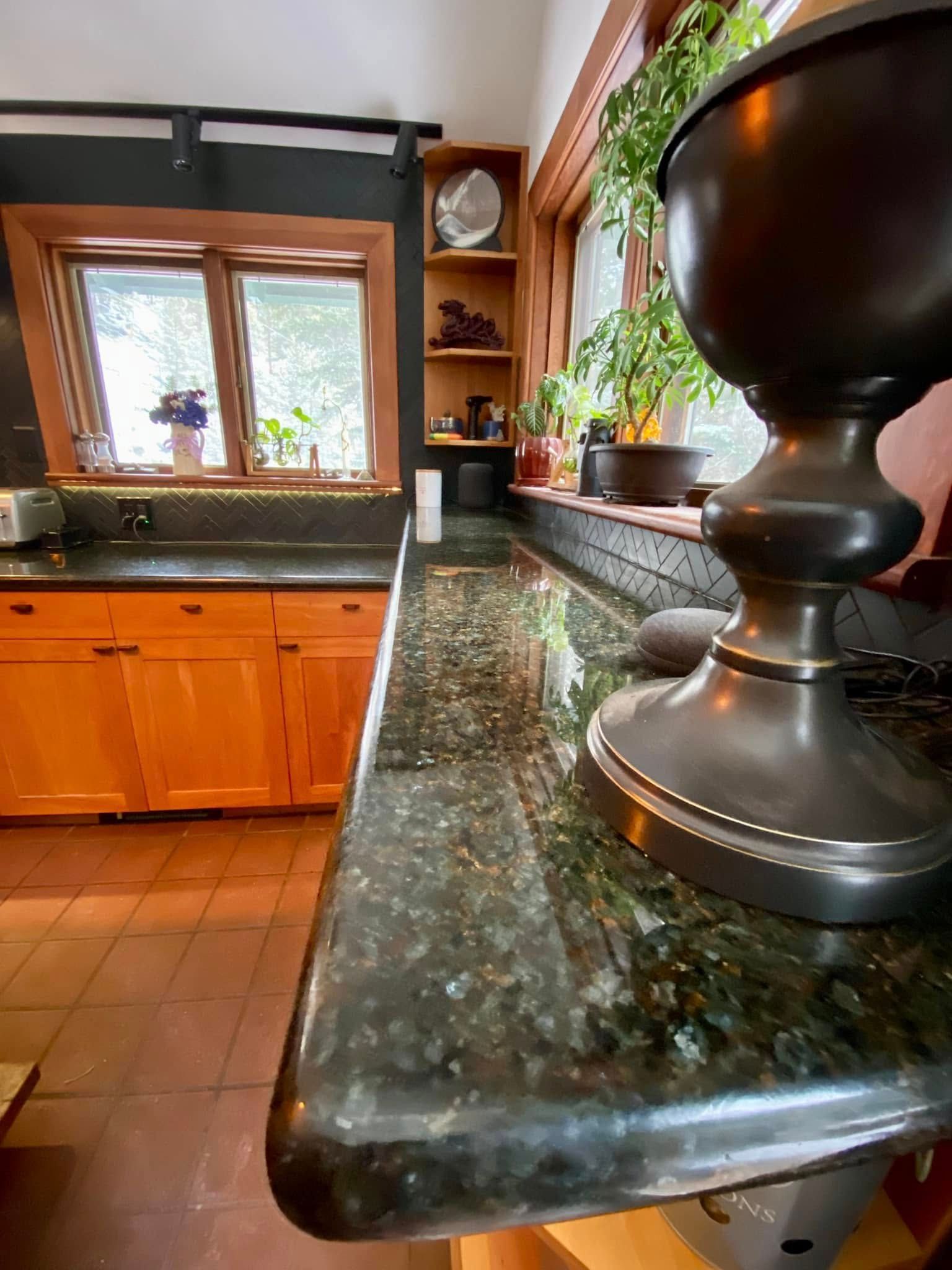 Kitchen countertop with dark green granite, wooden cabinets, window, and potted plant by the sink