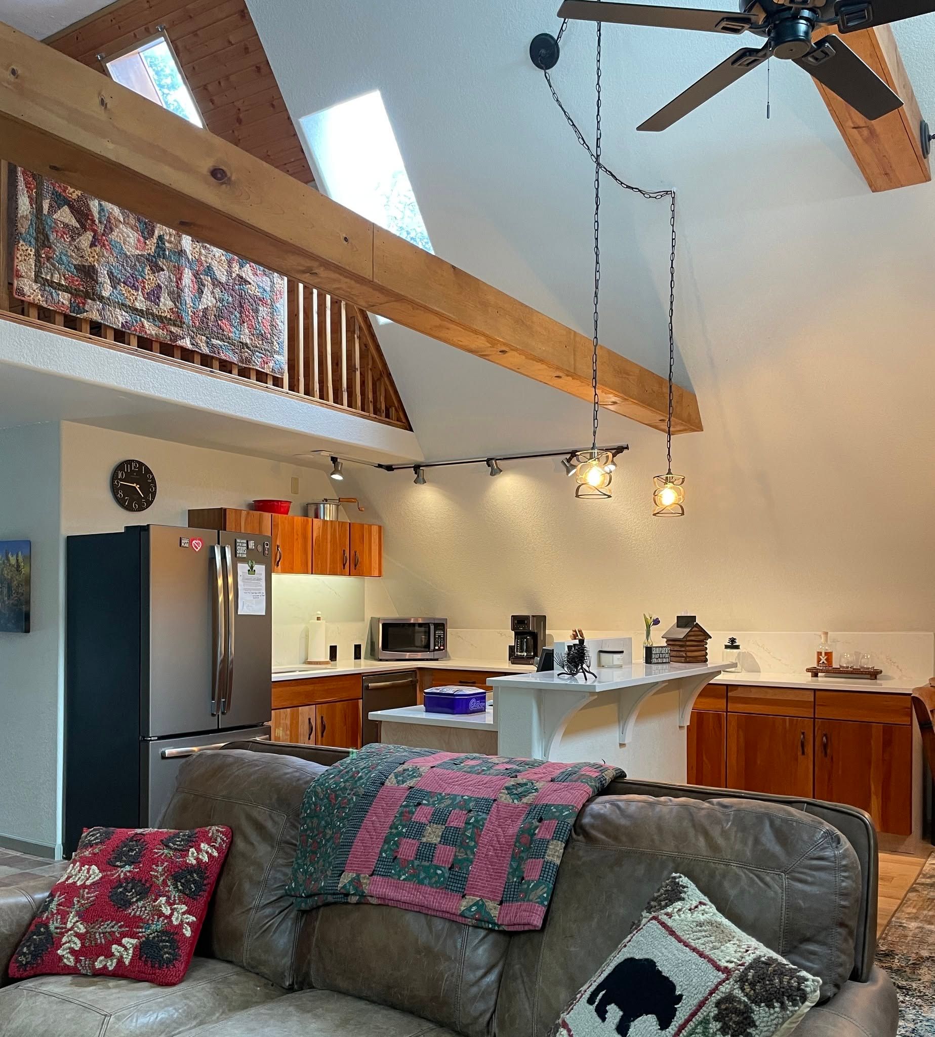 Bright open kitchen and living room with vaulted ceiling, wood beams, ceiling fan, and patterned sofa.