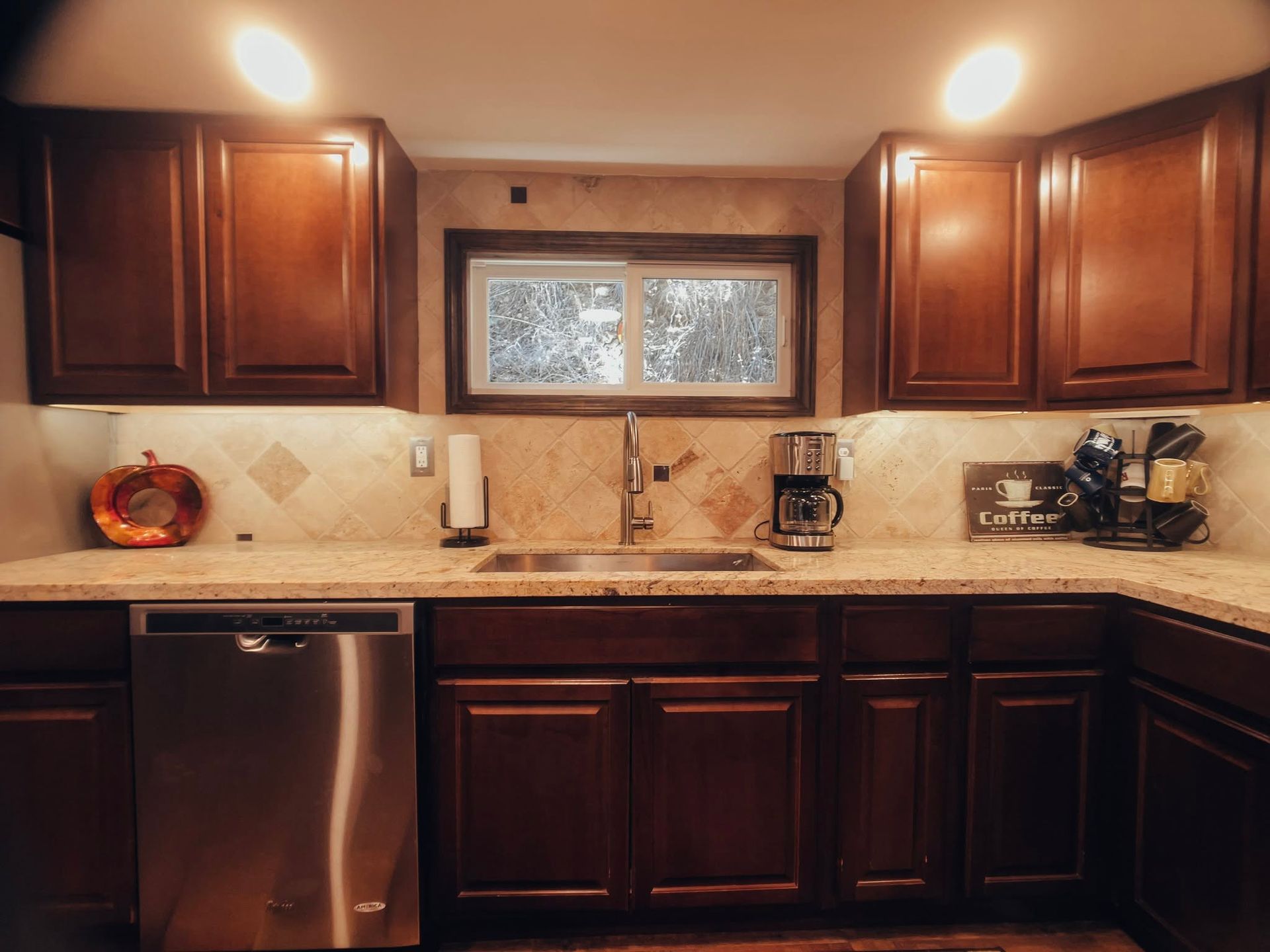 Warm kitchen with dark wood cabinets, stainless dishwasher, and a small frosted window above beige tile backsplash