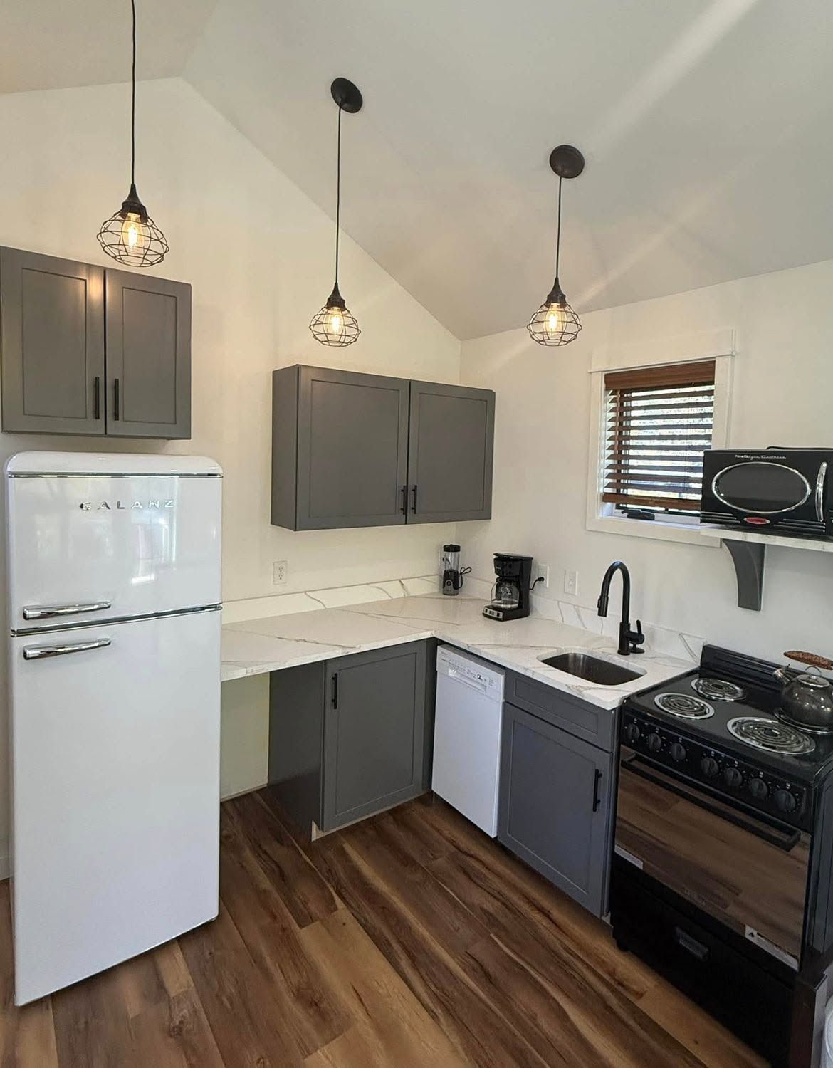 Compact kitchen with gray cabinets, white appliances, pendant lights, wood floor, and black stove and sink.
