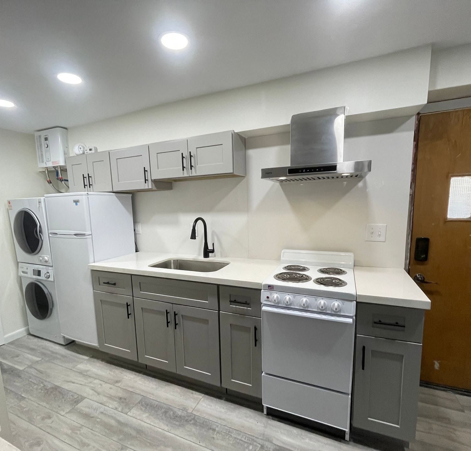 Small kitchen with gray cabinets, white countertops, sink, stove, range hood, and stacked washer-dryer.