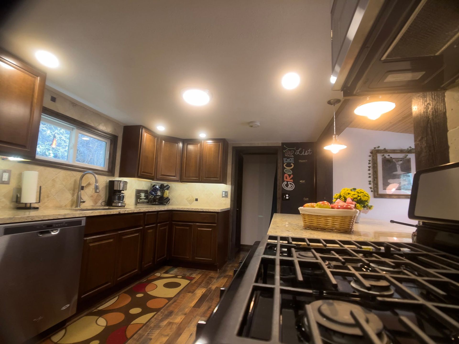 Modern kitchen with dark wood cabinets, stone counters, gas stove, and patterned rug under recessed lights