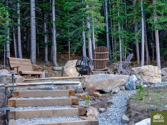 Wooded backyard fire pit area with stone steps, chairs, and large boulders