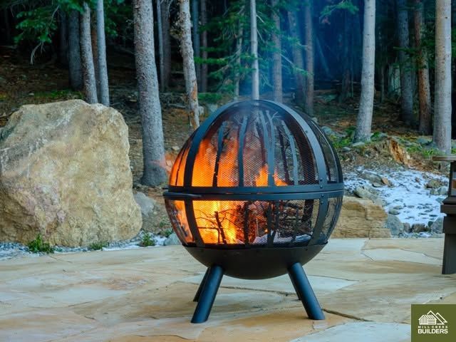 Fire burning in a metal globe fire pit outdoors on a stone patio, with trees and rocks in the background