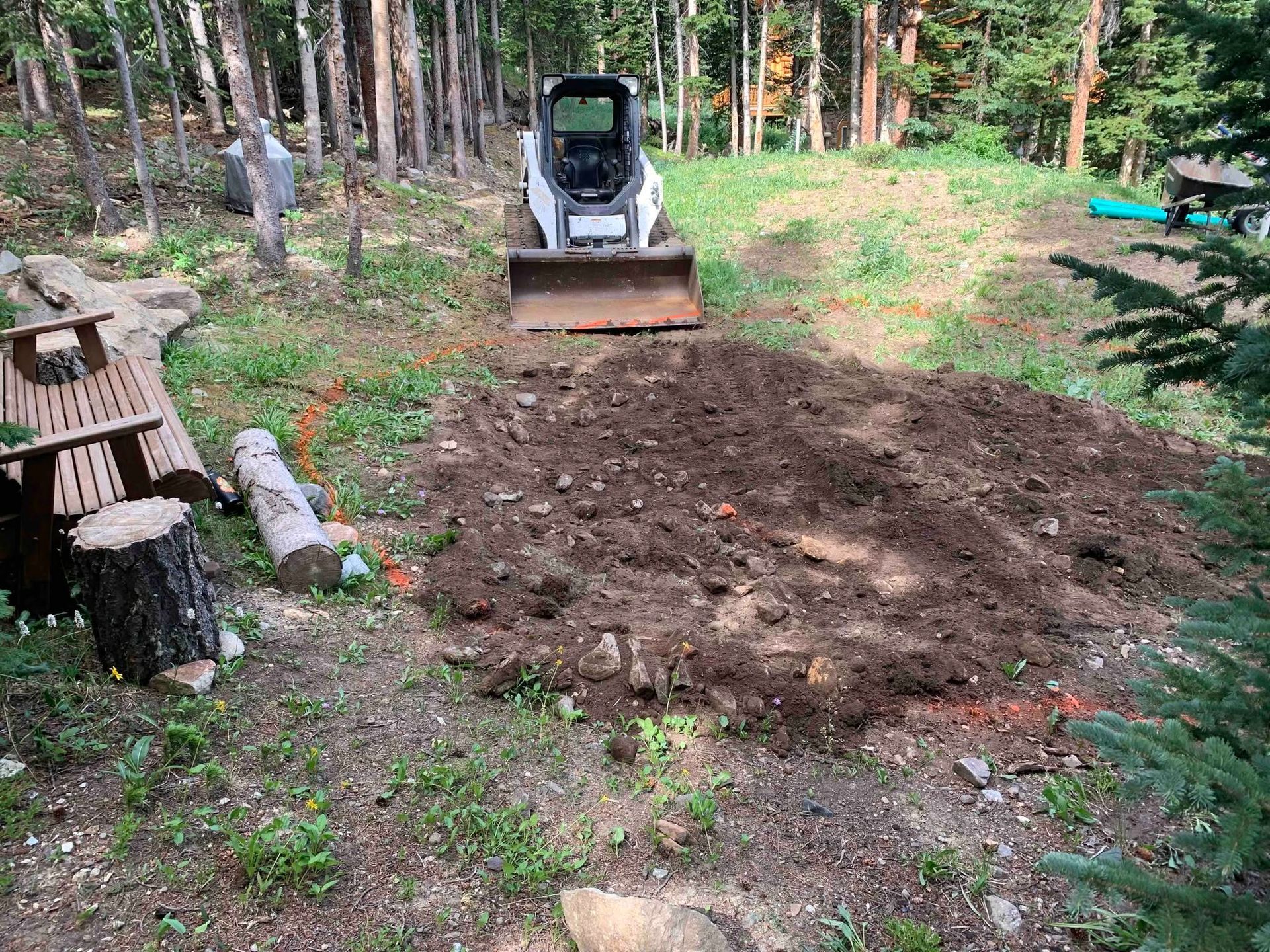 Freshly dug soil in a forest clearing with a small excavator and a wooden bench nearby