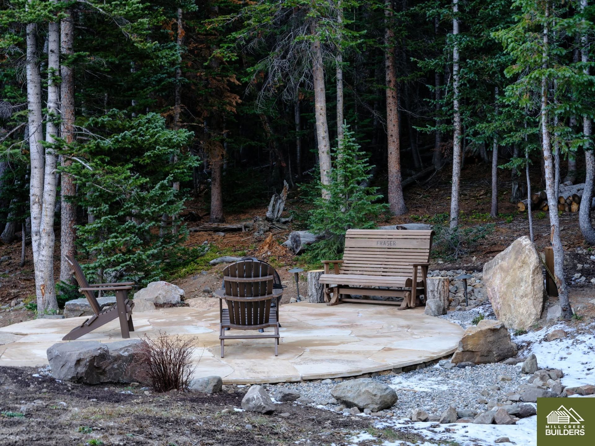 Wooden deck with chairs and benches around a fire pit in a wooded forest clearing
