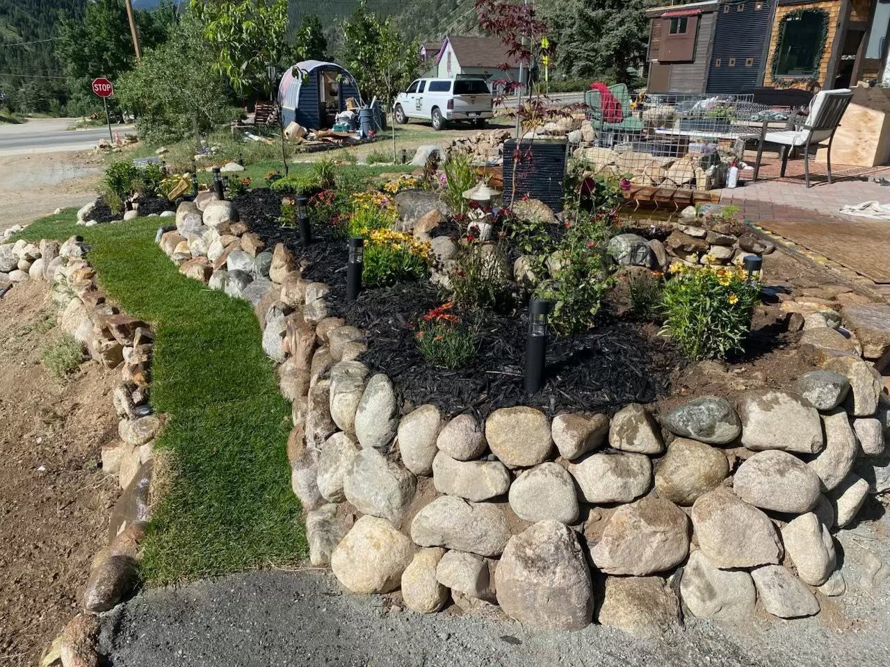 Stone-bordered garden beds with flowers and a grassy path in a sunny yard