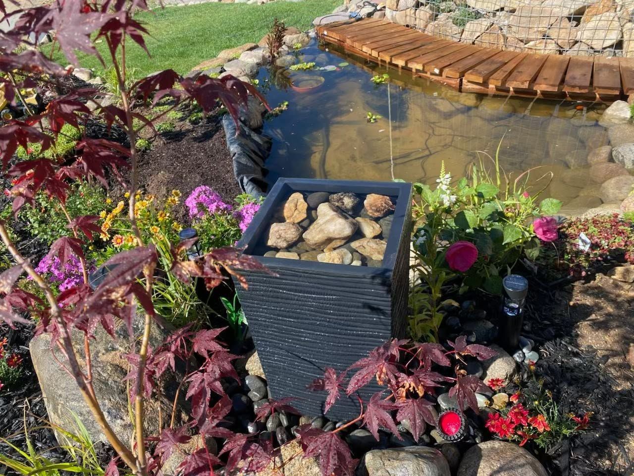 Garden pond with a black stone planter, colorful flowers, and a small wooden bridge in the background