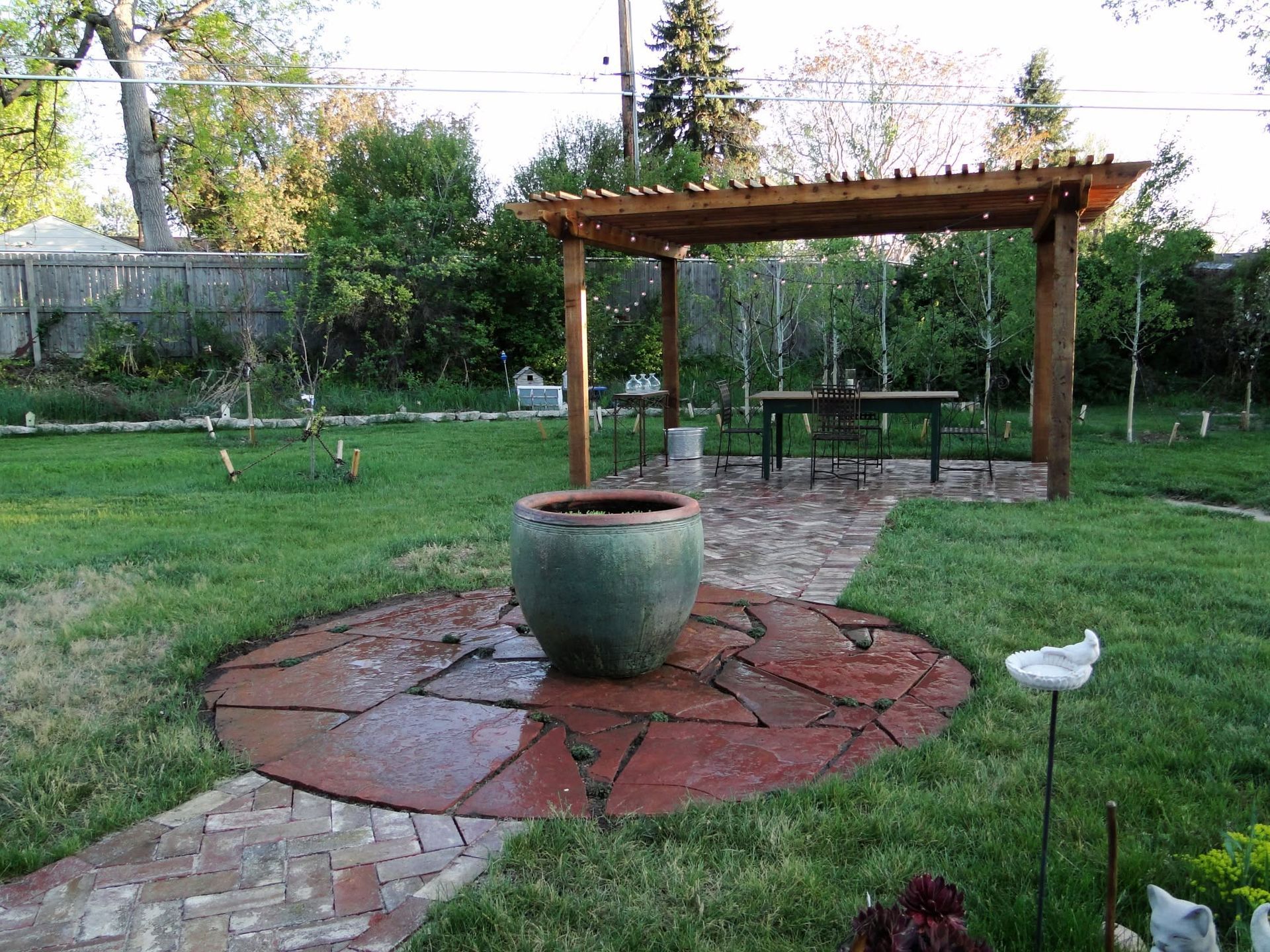 Backyard garden with a wooden pergola, large planter, and red stone patio surrounded by grass and trees