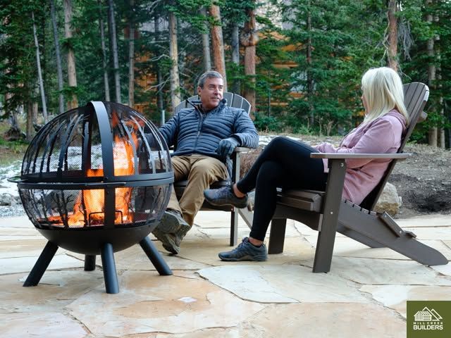 Two people sit by a round fire pit on a wooded patio, talking in folding lounge chairs.