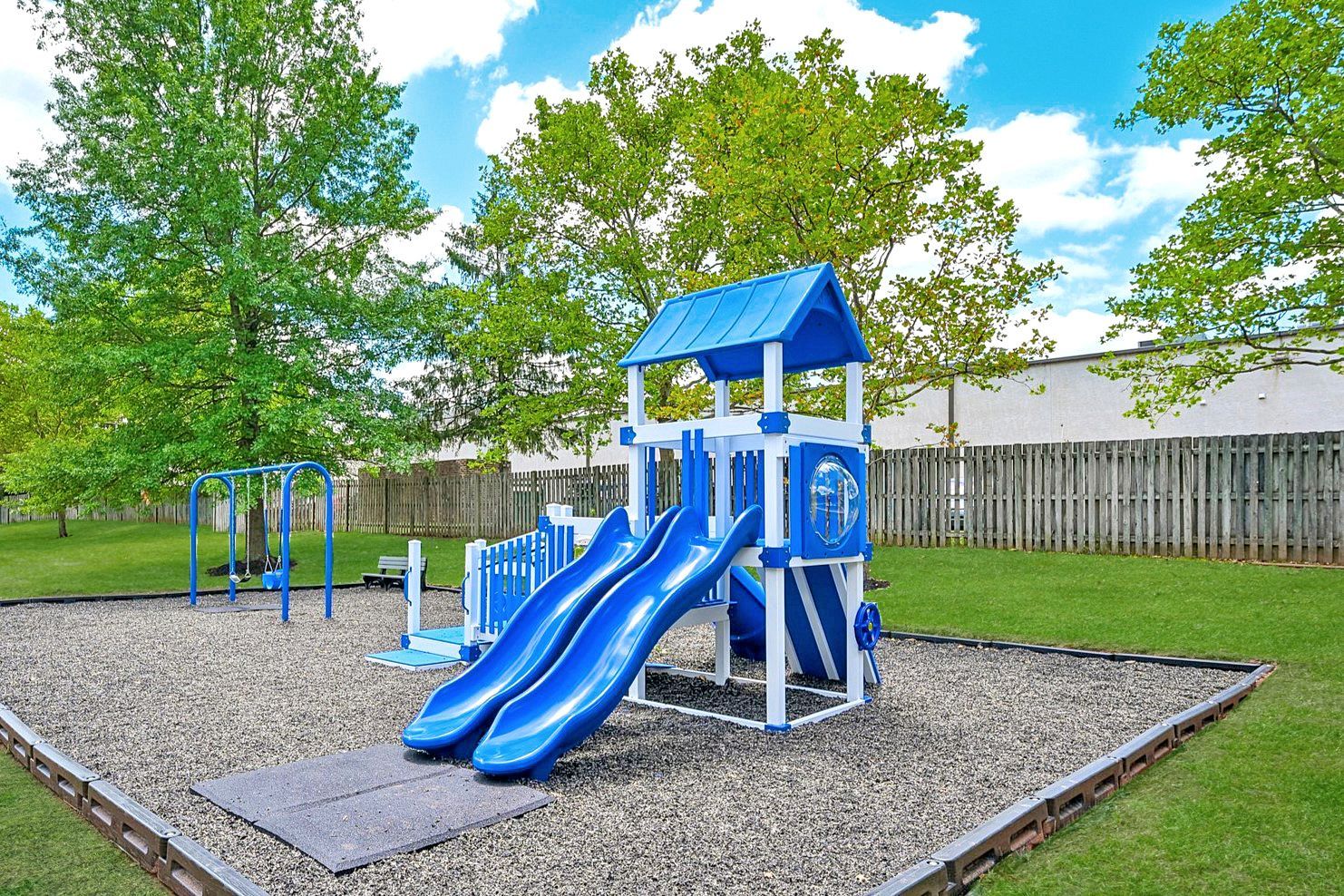 Blue playground slide and climbing structure in a gravel play area with trees and a wooden fence.