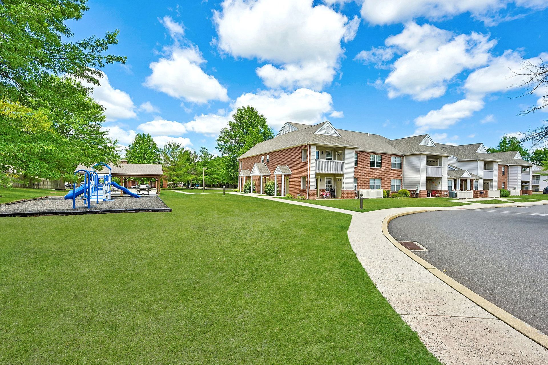 Apartment complex with brick buildings, green lawn, and a playground under a bright blue sky
