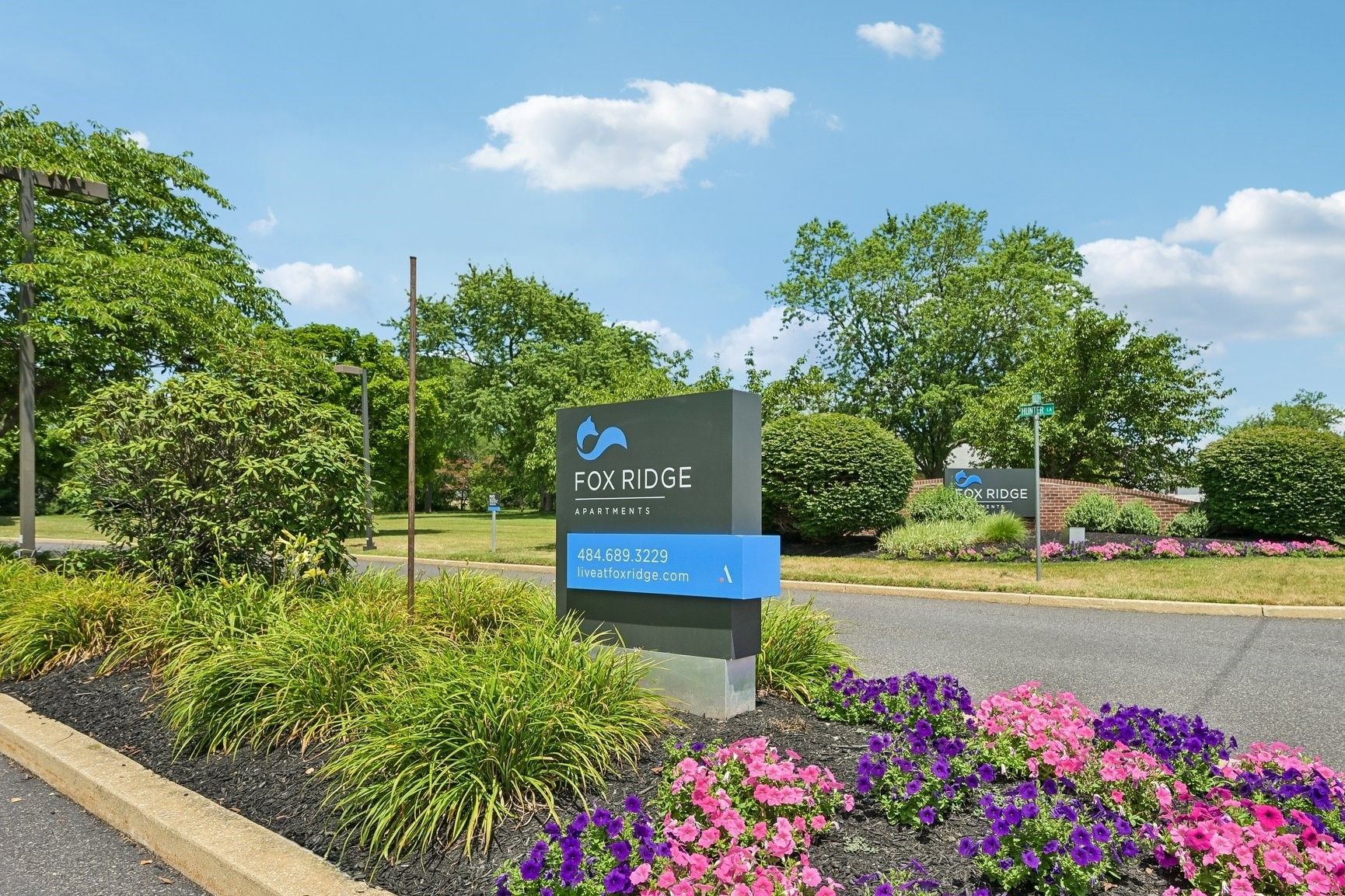 Outdoor sign at a landscaped entrance with flowers, trees, and a blue sky.