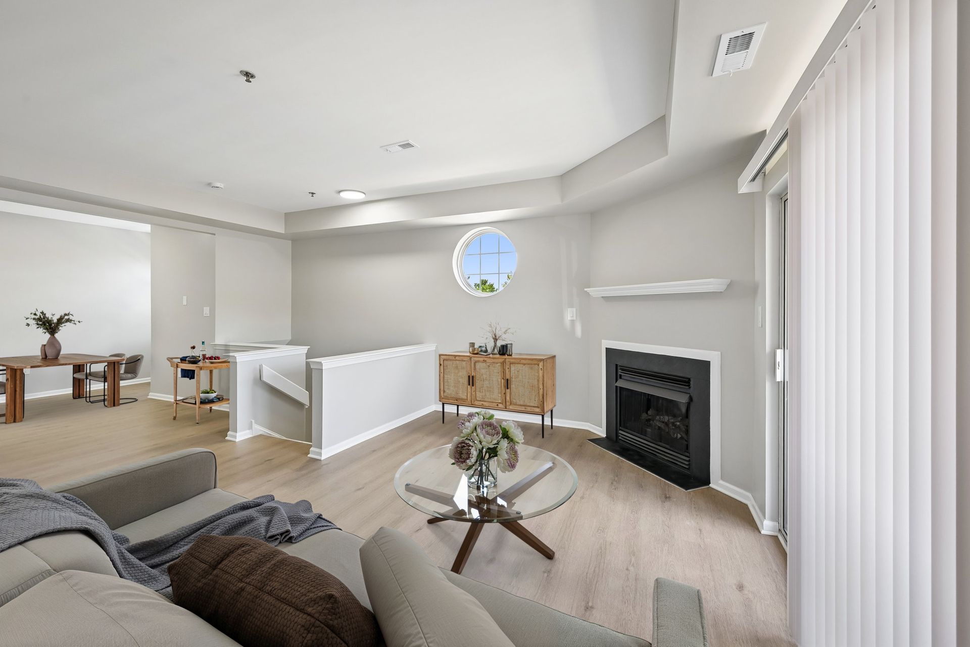 Bright modern living room with white walls, gray sofa, fireplace, and round window overlooking a dining area