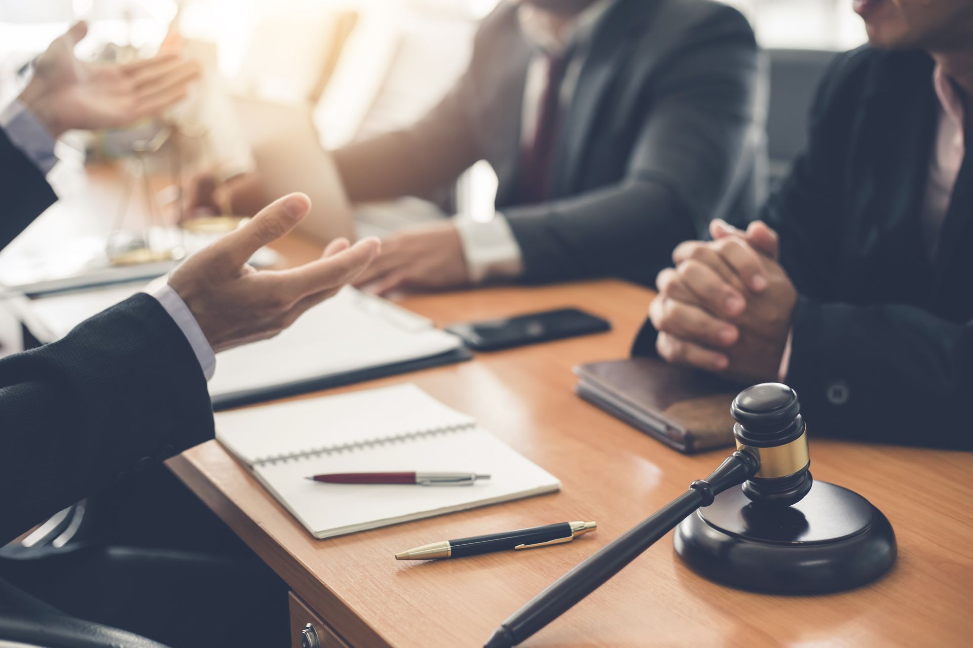Legal professionals in suits sit at a desk with notebooks, a pen, and a judge's gavel during a meeting. Legal professionals in suits sit at a desk with notebooks, a pen, and a judge's gavel during a meeting.