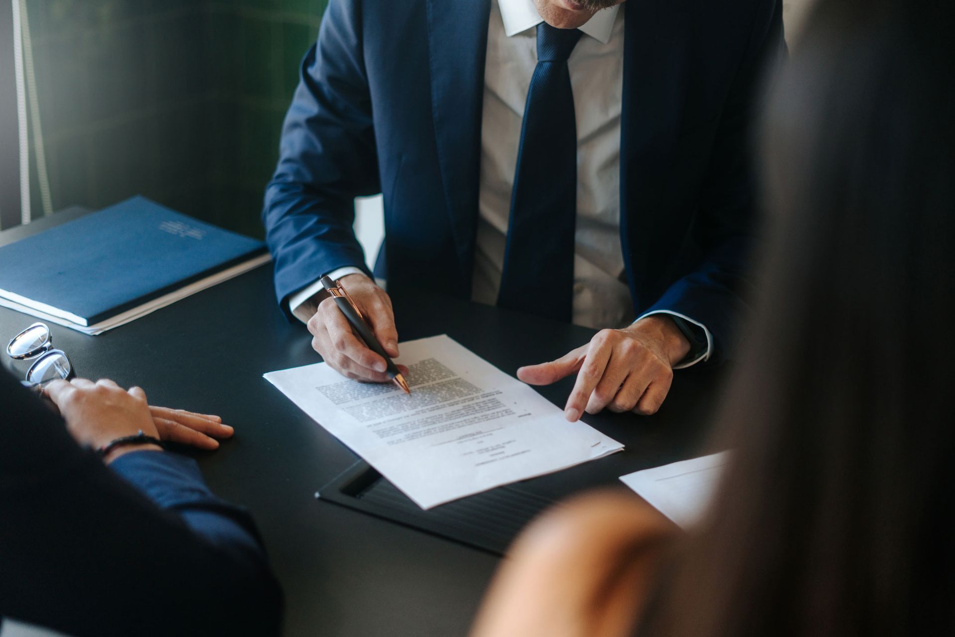 Person in a suit signing a document at a desk with two other people.