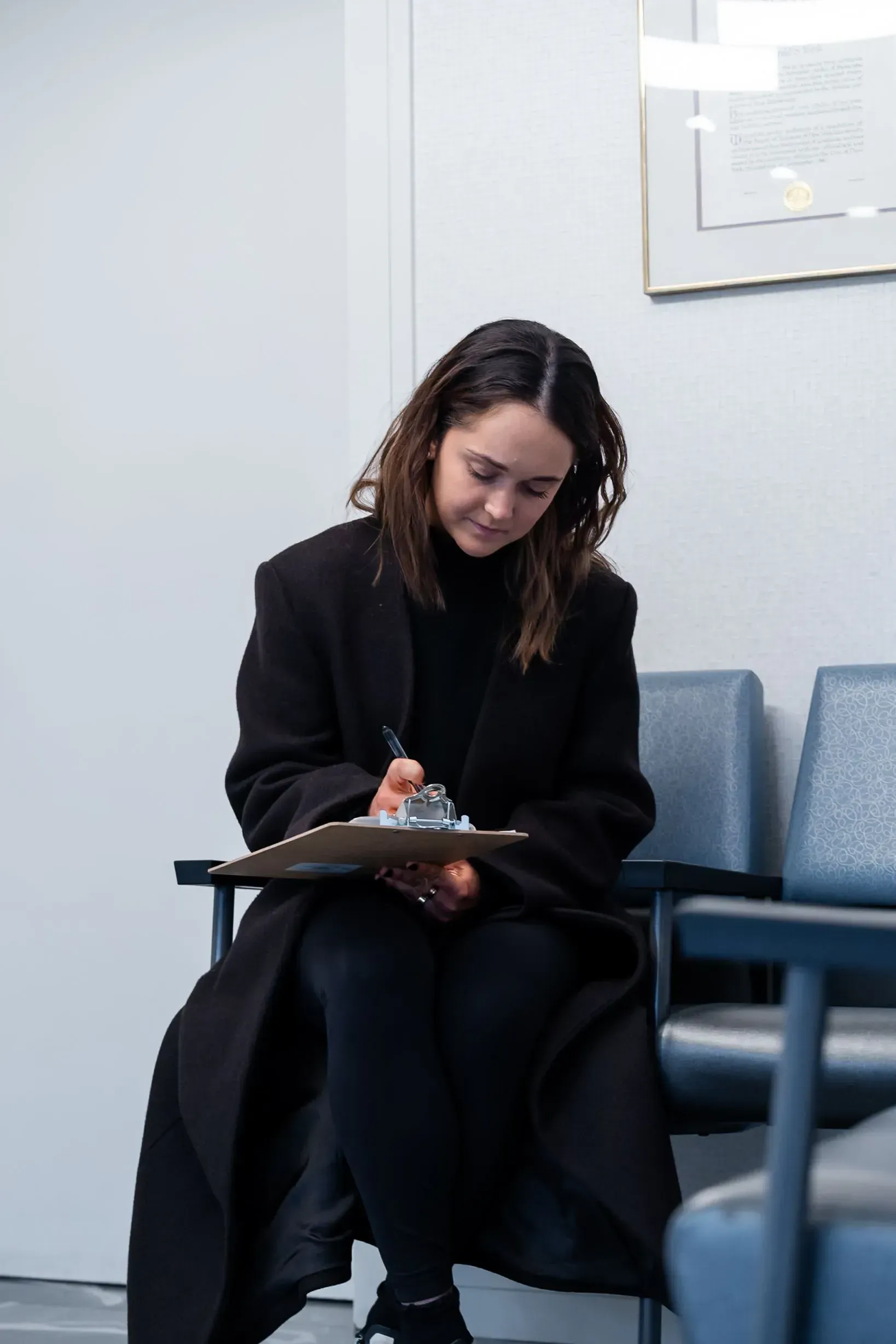 Person in a black coat writing on a clipboard while seated in a waiting area.