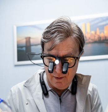 Doctor wearing surgical loupe glasses in a clinic, looking down in front of a framed cityscape.