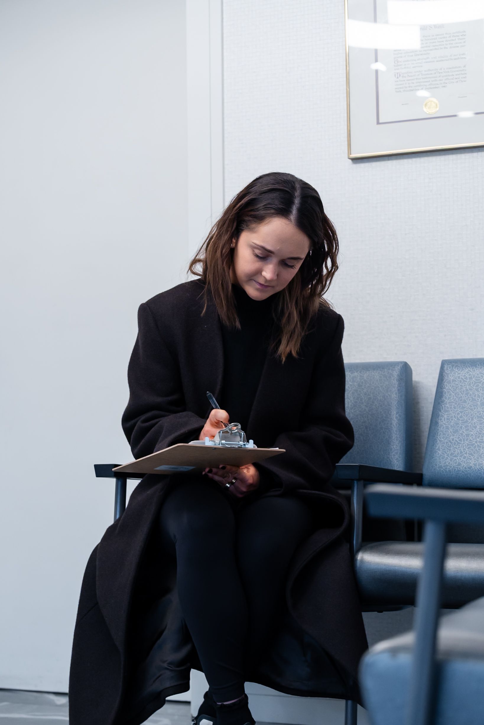 Person in a black coat writing on a clipboard while seated in a waiting area.