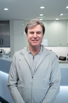 Man standing in a bright office kitchen, smiling in a light gray shirt.