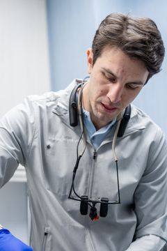 Doctor in a light gray coat with a stethoscope, looking down while examining a patient in a clinical setting.
