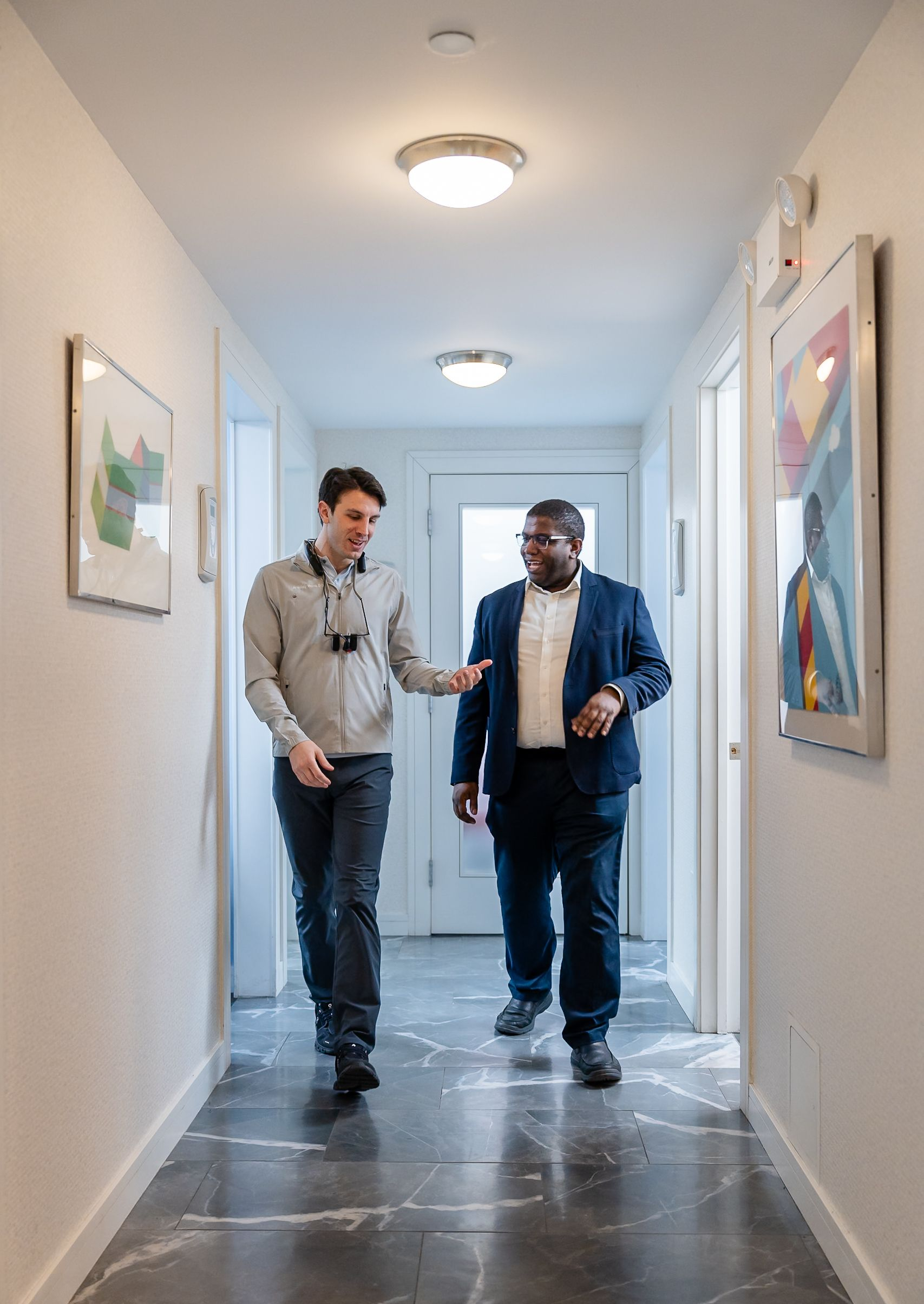 Two men talking in a bright hallway, one holding a tablet and the other gesturing while walking