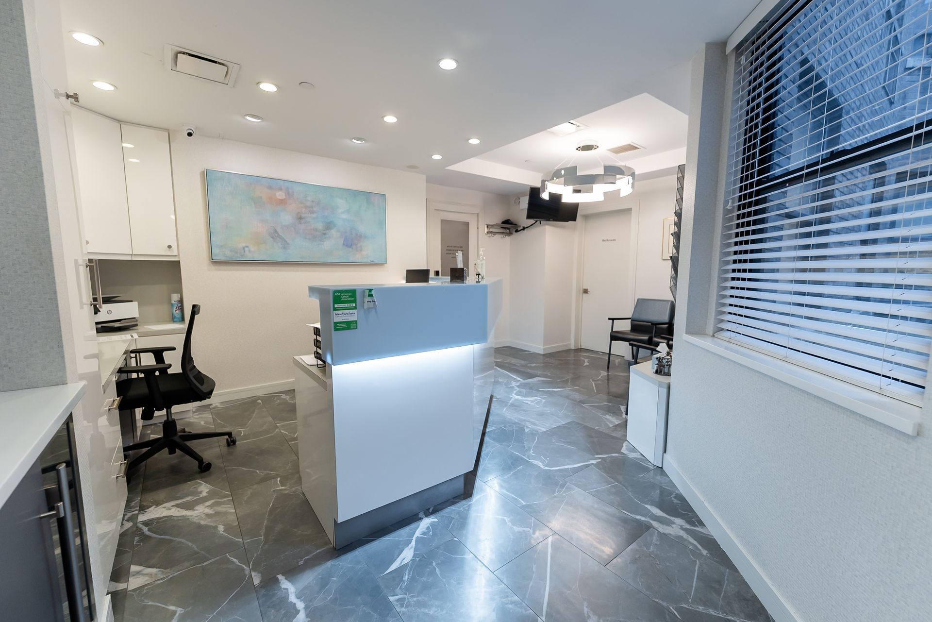 Modern office reception area with white walls, marble floor, blue-accented desk, chairs, and wall art.