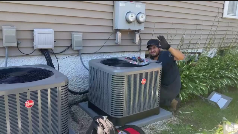 Technician beside two outdoor air conditioning units, waving in front of a house siding wall.