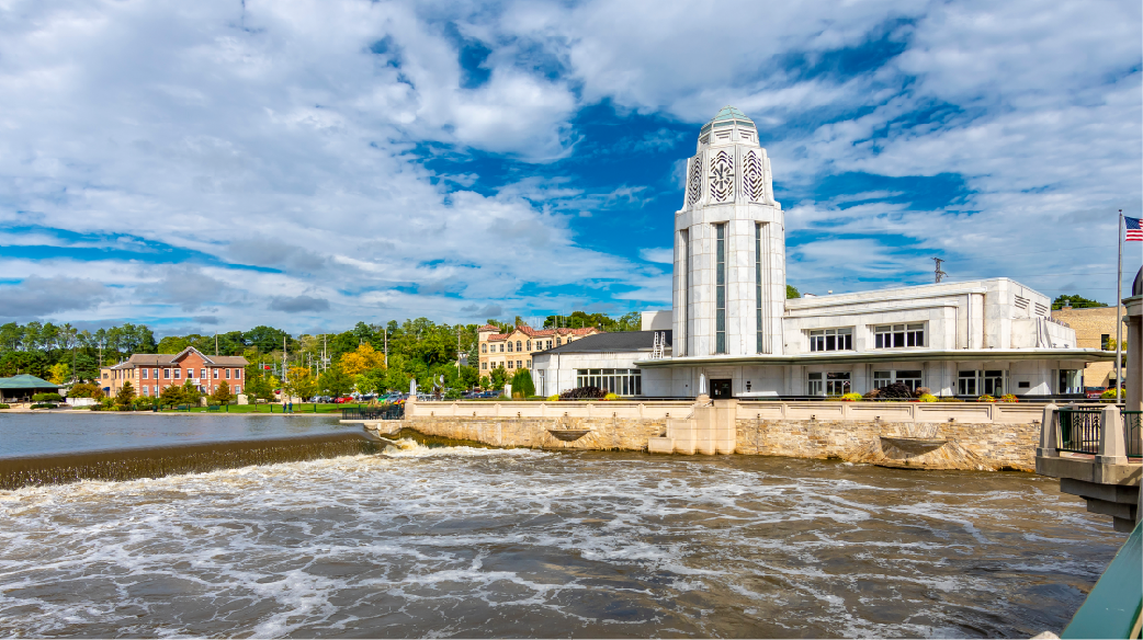 Tall white riverside building by a waterfall under a blue sky with scattered clouds
