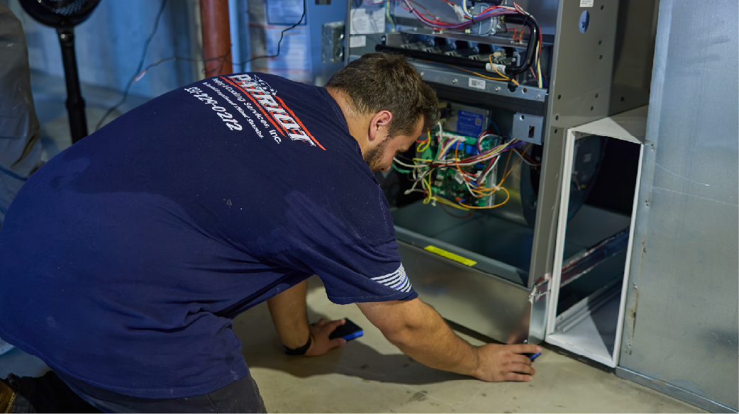 Technician kneeling beside an open electrical panel, inspecting wires in a utility room.