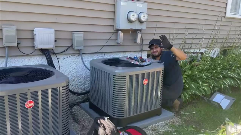Technician kneeling beside outdoor air conditioners, waving next to a house wall.