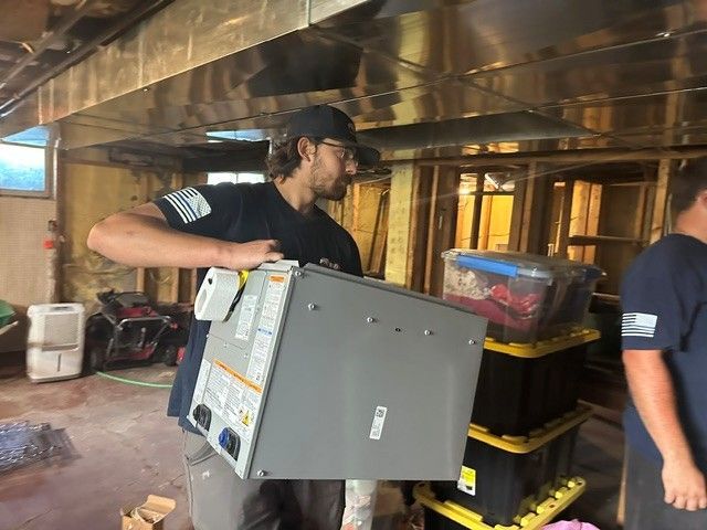 Worker carrying a large gray appliance through a cluttered workshop or garage