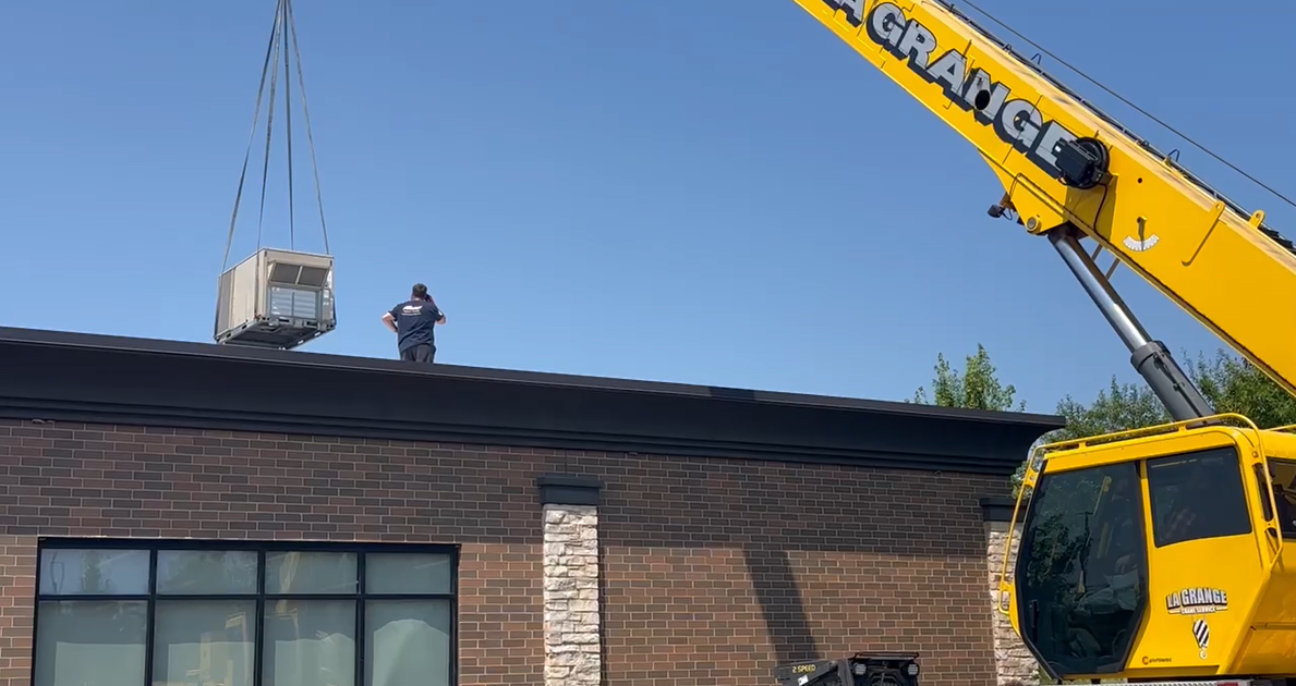 Crane lifting an HVAC unit onto a brick building roof under a clear blue sky
