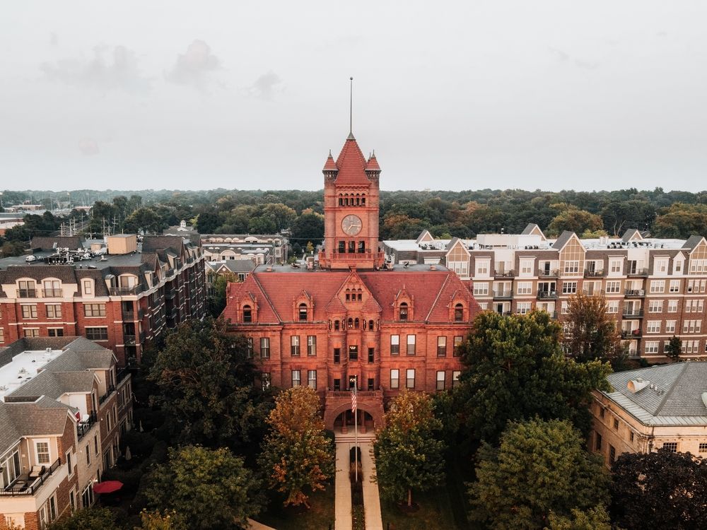 A red-brick historic building with a central tower, framed by trees and row houses under a cloudy sky.
