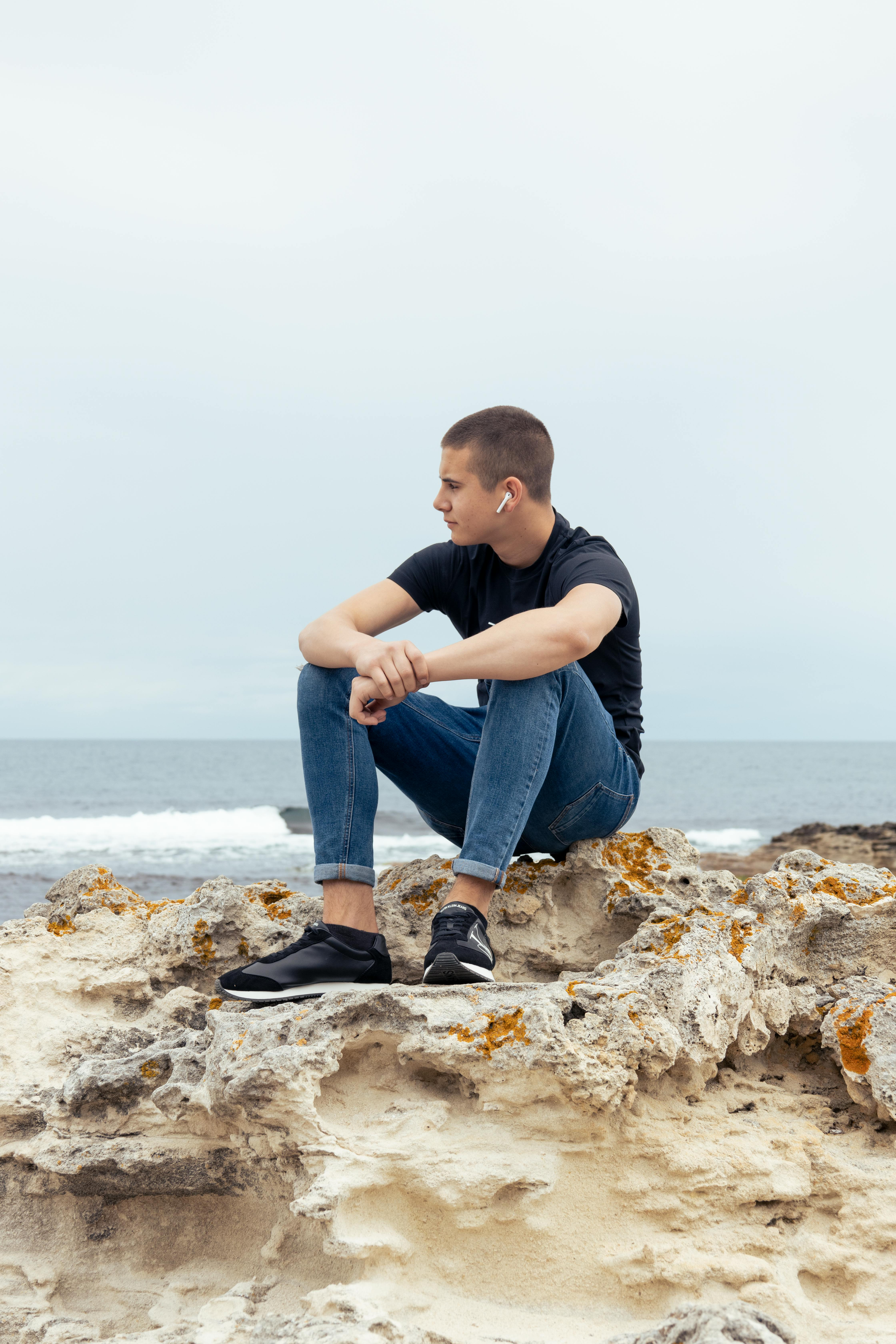 Man zit op rotsen bij de zee en kijkt weg. Hij draagt een spijkerbroek, een zwart shirt en sneakers.