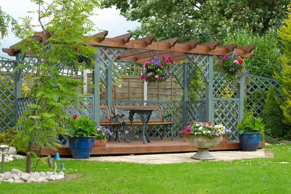 Garden patio with wooden pergola, table and chairs, hanging flowers, and potted plants on a lawn backdrop