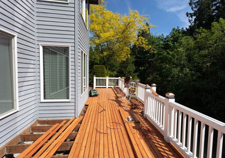Wooden deck beside a gray house, with white railing and autumn trees under a blue sky.