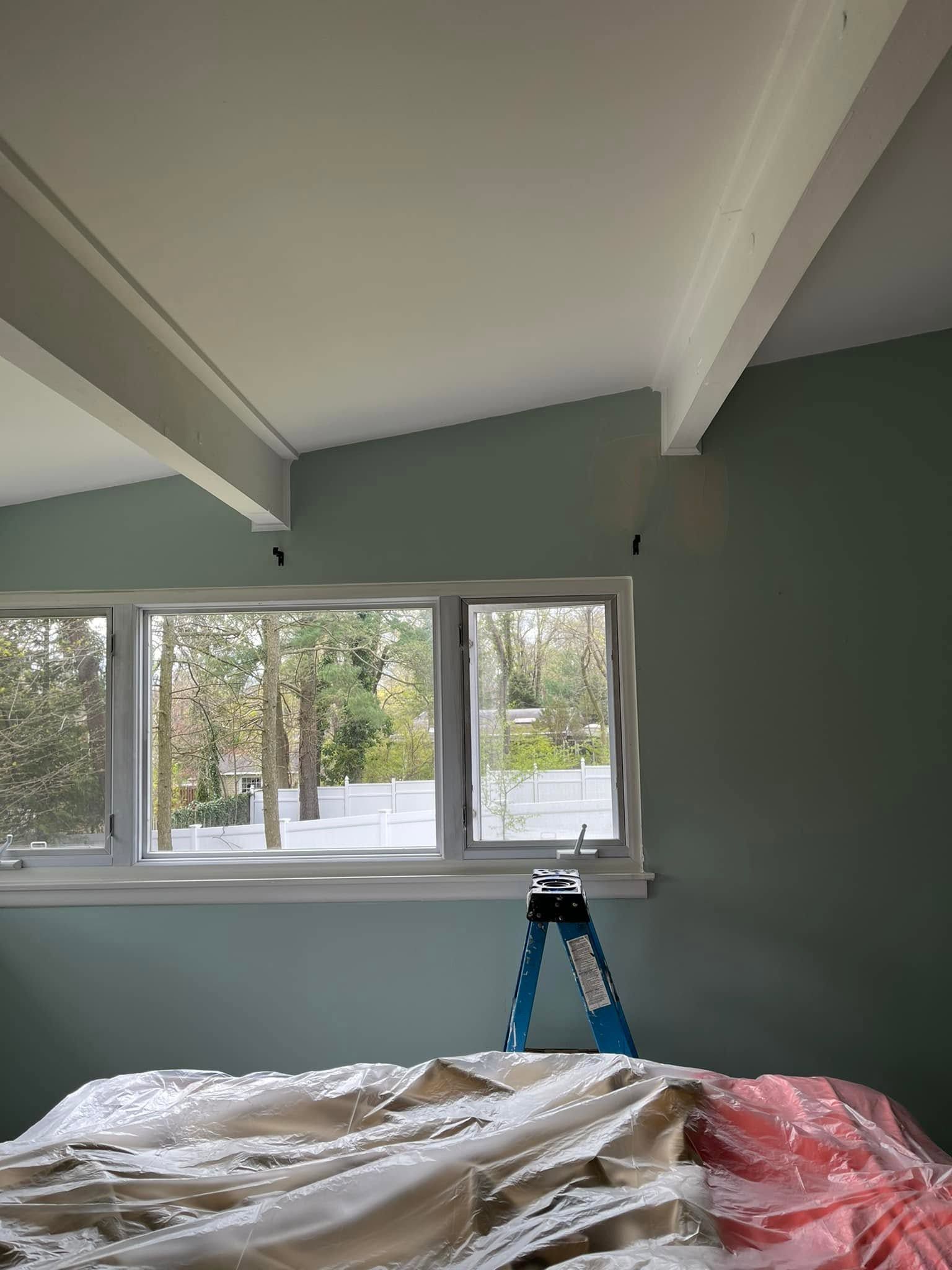 Partially renovated room with green walls, exposed ceiling beams, windows, and a ladder over covered furniture
