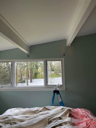 Living room with white ceiling beams, green walls, window view, ladder, and covered furniture under renovation